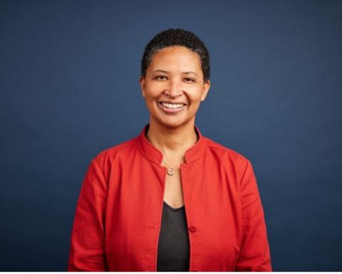 Danielle Allen stands in front of a blue background wearing a red jacket and black top. She has short black hair and is smiling.