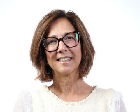 Tracy Battaglia has a brown bob and eyeglasses. She is wearing a white blouse and has her arms crossed.