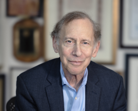Bob Langer is wearing a dark blue blazer and light blue shirt. He is sitting in a chair with his hands clasped. There are frames on the wall behind him.