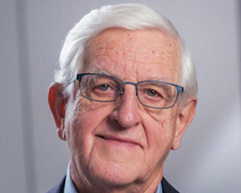Joseph Martin has short white hair and is wearing a blue shirt with a dark blue blazer. He also has blue-framed glasses.
