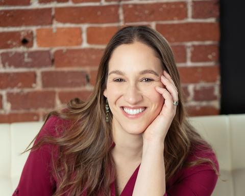 Rachel Fish is sitting on a white couch with a brick wall background. She is wearing a maroon blouse and blue jeans.