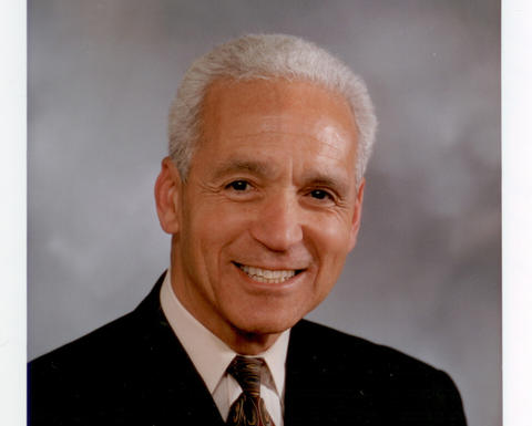 Gus White has short grey hair and is wearing a black blazer, white shirt, and brown tie.