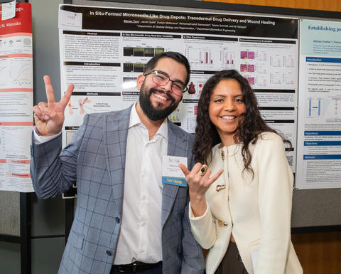 Two presenters at NESS make a peace sign and shaka sign respectively while standing in front of some research posters.