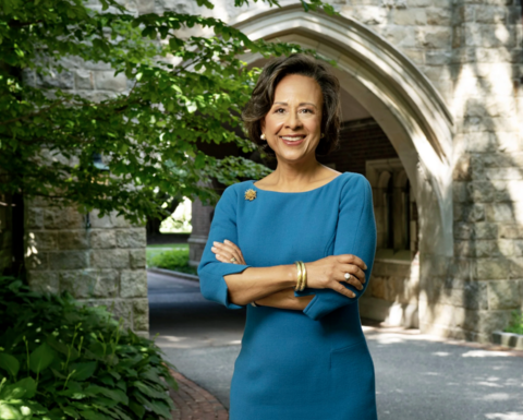 Paula Johnson is wearing a teal dress and gold accessories. She is standing outside a stone arch with greenery on either side. 