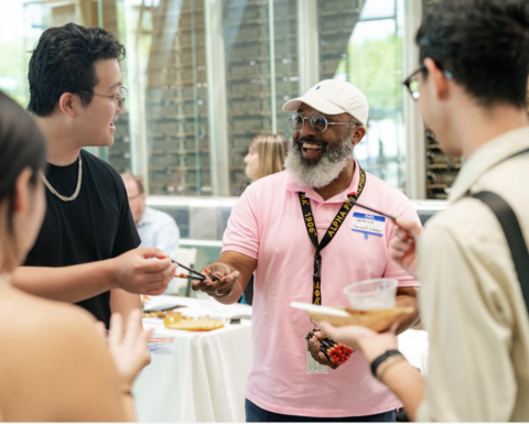 A group of people stand in a circle while a man in a pink polo in the middle passes out pencils.