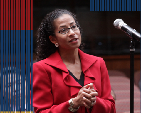 A women with long curly hair and glasses speaks into a microphone. She is wearing a ruffled red blazer.