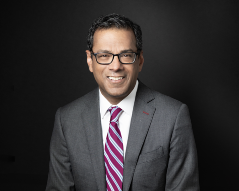 Atul Gawande is posing with his hand across his lap. He is wearing a gray suit, striped tie, and glasses.