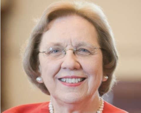 Barbara McNeil has short blonde hair and is wearing glass, pearl earrings, and a red blazer.