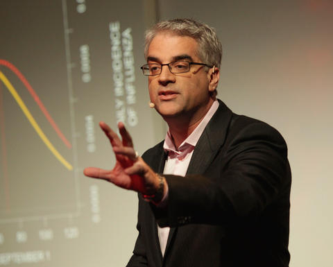 Nicholas Christakis has short gray hair and glasses. He is gesturing away from himself as he stands in front of a dark screen showing a graph.