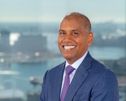 Mohan Suntha is a bald man wearing a blue suit and purple tie. A body of water and some buildings are visible behind him.